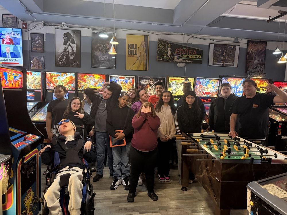 students standing in an arcade with arcade games behind them and a foosball table in front of them