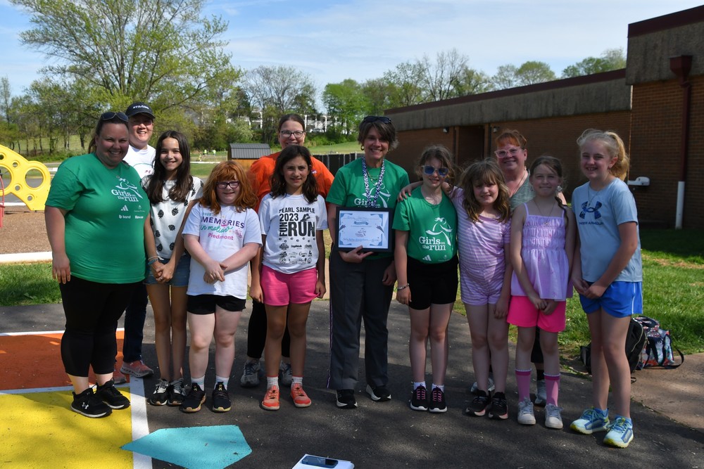A group of elementary-aged girls and adult coaches stand together outdoors on a school playground, smiling for a photo. Several of the adults are wearing green “Girls on the Run” shirts. One woman in the center holds a framed certificate, likely recognizing a milestone achievement. The group stands on a paved area with colorful playground markings, with a school building, grass, and trees visible in the background on a sunny day.