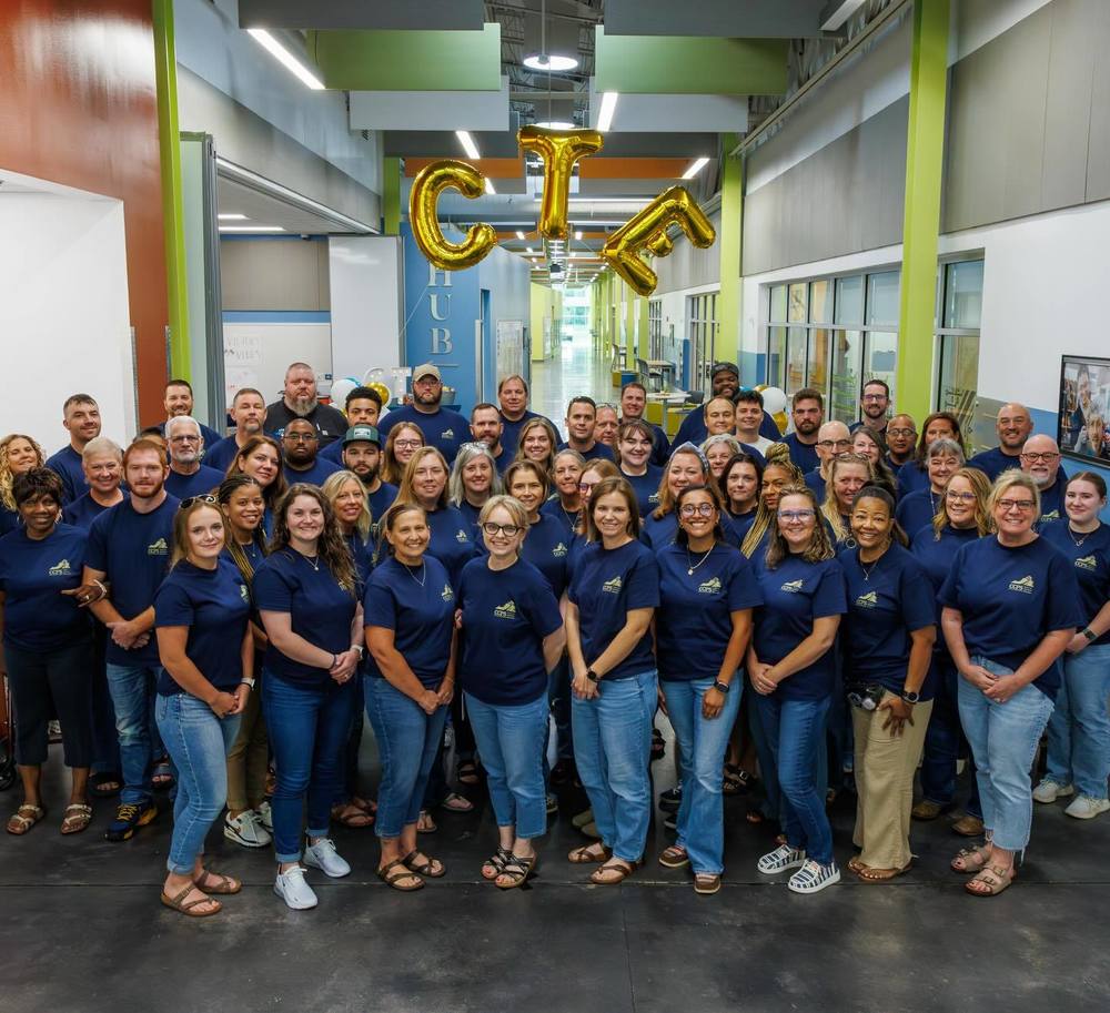 large group os teachers wearing matching blue shirts and jeans under gold balloons of the letters C T E 