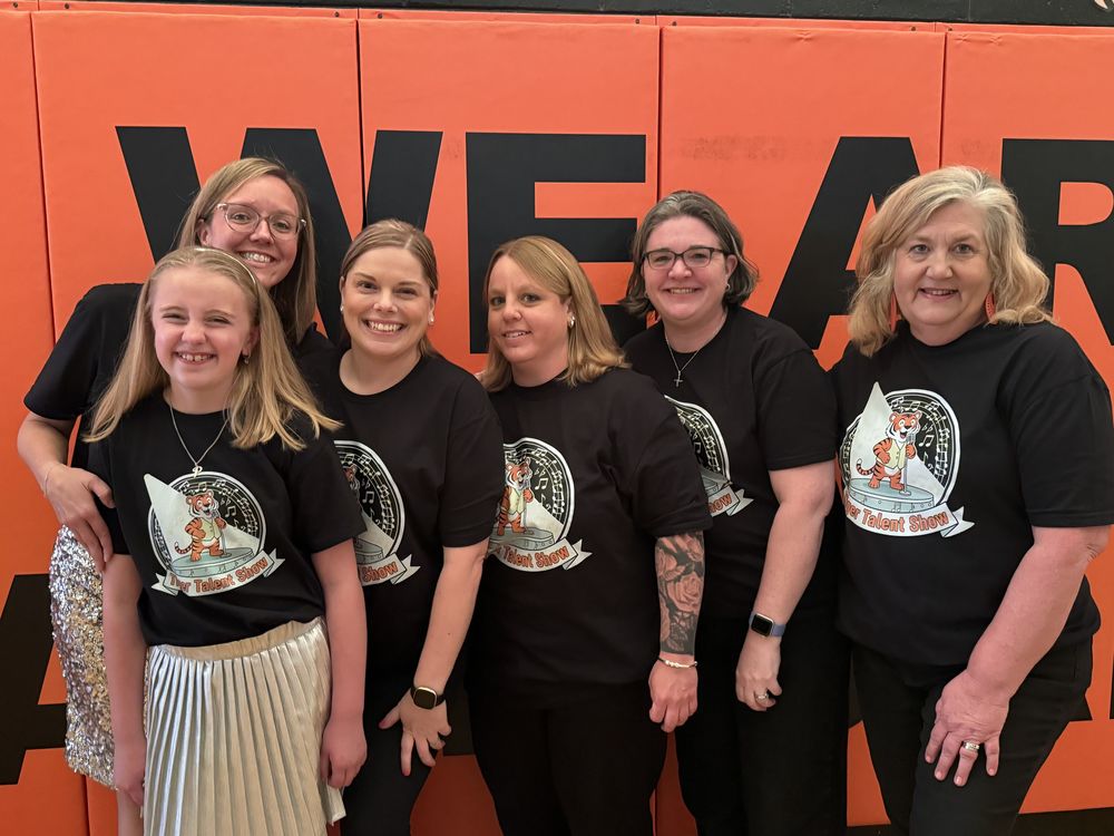 A group of six females—five adults and one young girl—stand smiling together in front of an orange wall with large black lettering. They are all wearing matching black T-shirts featuring a tiger graphic and the words “Talent Show.”