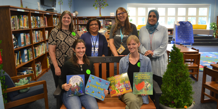 Image contains 4 adults standing behind a bench, two adults sitting on a bench holding books next to a plant in the school library.  The bench reads  "In loving memory of our favorite teacher, Evelyn Sawyer, who taught us the true, the good, and the beautiful."