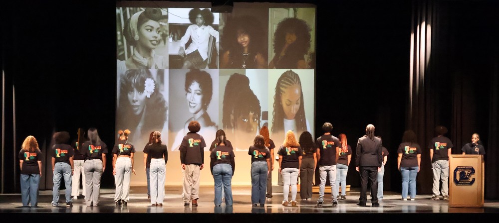 students standing on a stage with their backs to the audience wearing matching black history month tshirts with images of black women on the screen behind them
