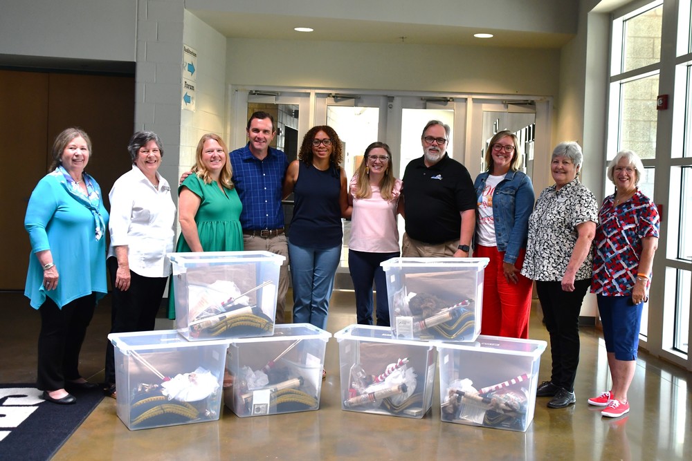 Members of the local Daughters of the American Revolution chapter and Culpeper County Public Schools staff stand together behind six large clear plastic tubs filled with colonial-era educational materials. The group is gathered in a school hallway for the donation presentation.