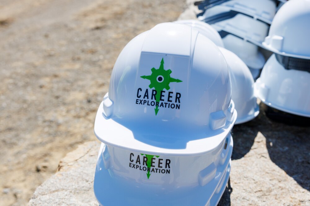 Close-up of white hard hats featuring the green and black “Career Exploration” logo, stacked together at a construction site.