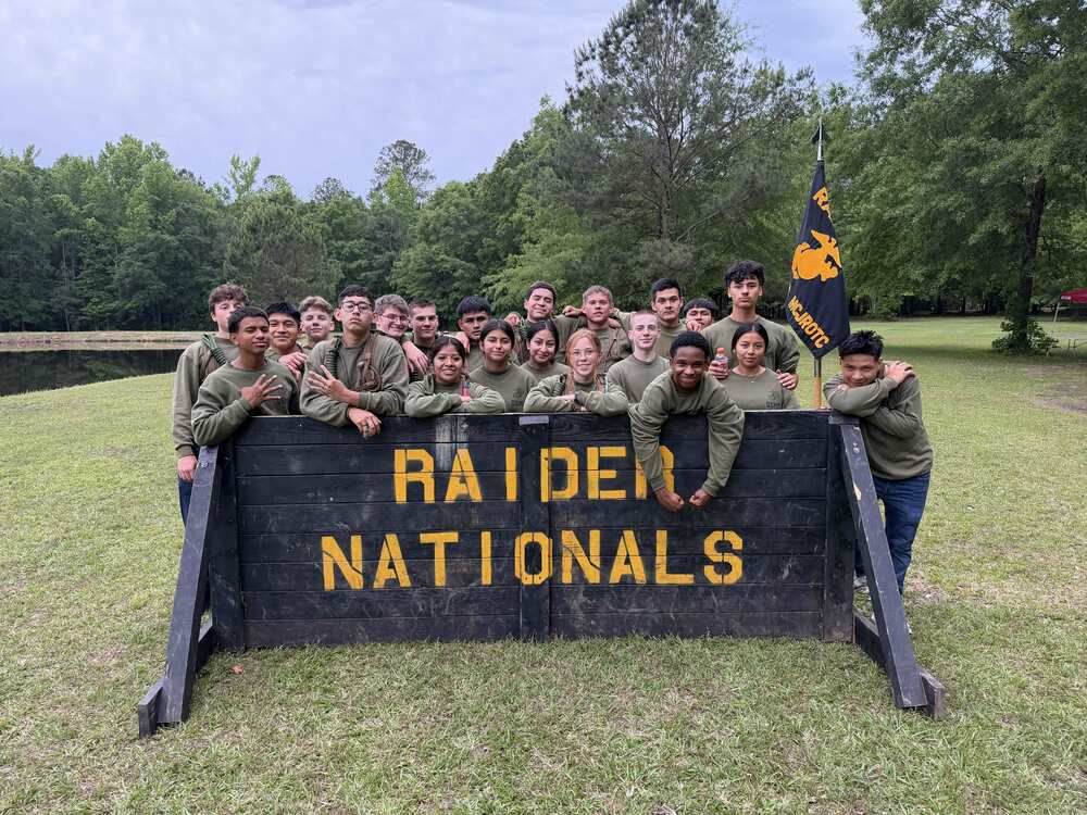 large group of junior rotc students in army green sweatshirts behind a black sign that says raiders nationals in yellow letters