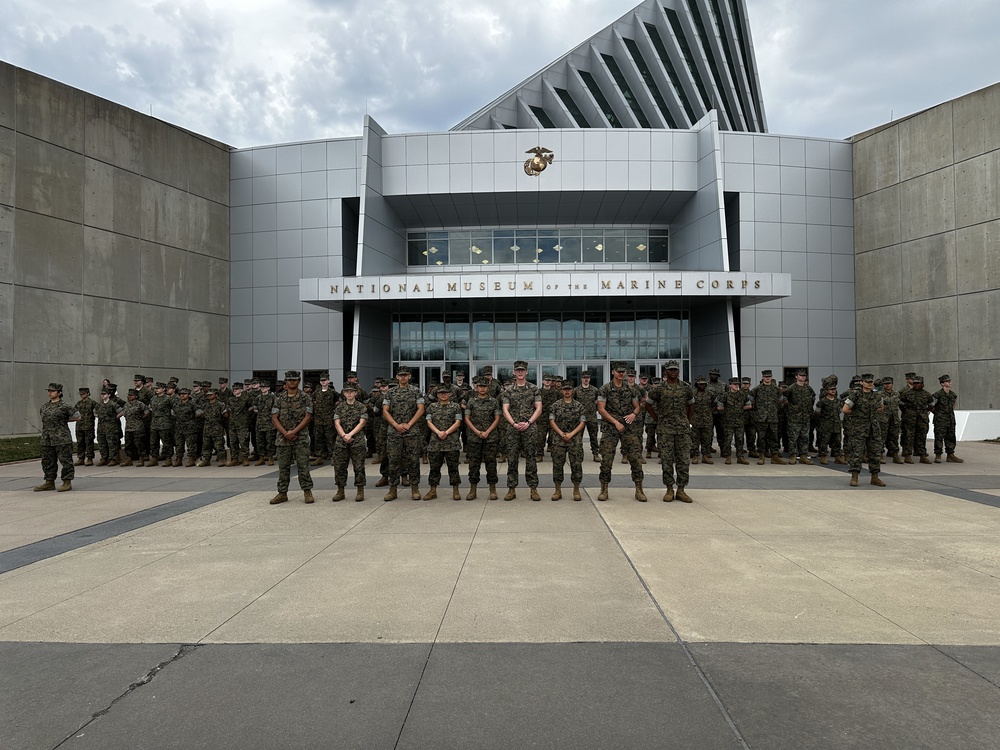 A large group of JROTC cadets in camouflage uniforms stands in formation in front of the National Museum of the Marine Corps, posing together beneath the building’s distinctive angular architecture.