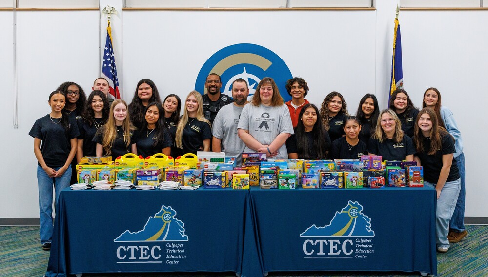 group of students in matching blue tshirts standing behind 2 tables with 2 adults in gray shirts in the midde, the tables are holding over 100 lego sets, and the tablecloths have the CTEC logo on them