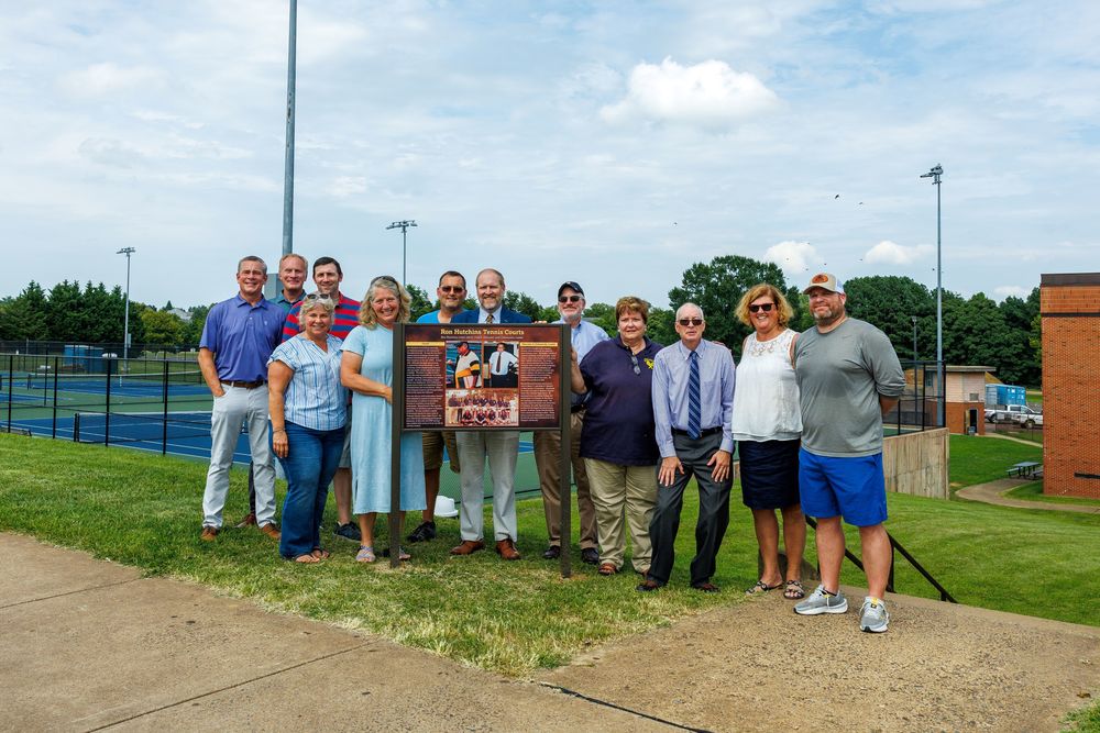 A group of fourteen people stands together outdoors beside a large commemorative plaque that reads "Ron Hutchins Tennis Courts." The plaque features photos of Coach Hutchins and his tennis teams, along with a written tribute. The group includes men and women of varying ages, smiling and posing in front of the newly dedicated tennis courts, which are visible in the background with blue and green playing surfaces. The setting is on the campus of Culpeper County High School, with a brick building to the right and light poles and trees in the distance under a partly cloudy sky.