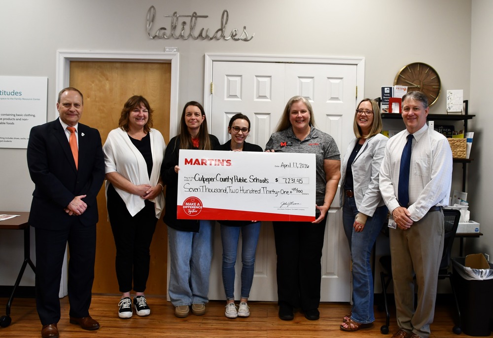 A group of seven adults stand indoors in front of double white doors beneath a wall sign reading “latitudes.” Three women in the center hold a large ceremonial check from MARTIN’S made out to Culpeper County Public Schools for $7,231.45, dated April 17, 2020. The group is smiling and dressed in business-casual attire, with one man in a suit on the far left and another on the far right. A wooden door, wall clock, shelving unit, and office items are visible in the background, suggesting a community resource or office setting.
