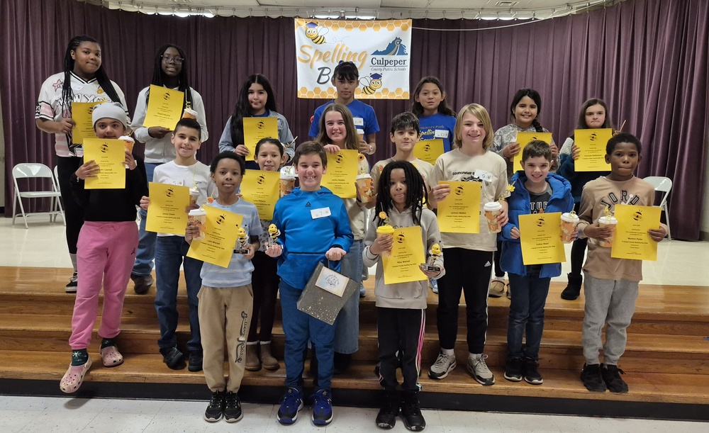 group of elementary students on a school stage holding certificates. there is a sign above them that says CCPS Spelling Bee in front of a purple curtain