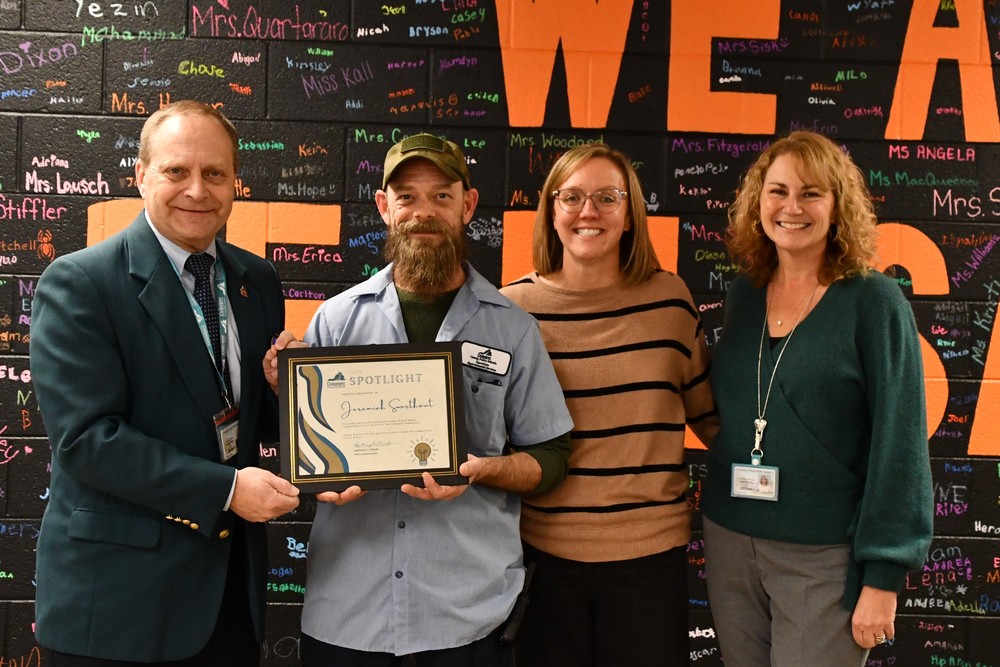 four people standing in front of a black and orange decorated wall, with the superintendent on the left, and  PSES custodian next to him both holding a CCPS Spotlight certificate, the principal and the HR Director are standing next to them on the right