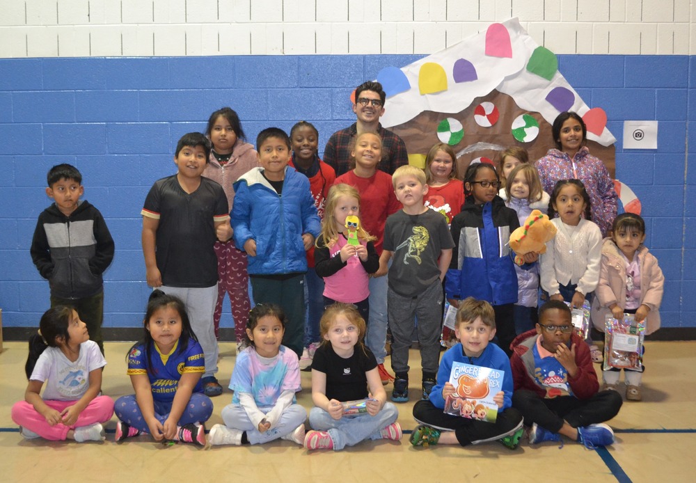 group of students in school gym with author in the back row.  there is a paper gingerbread house on the wall behind them