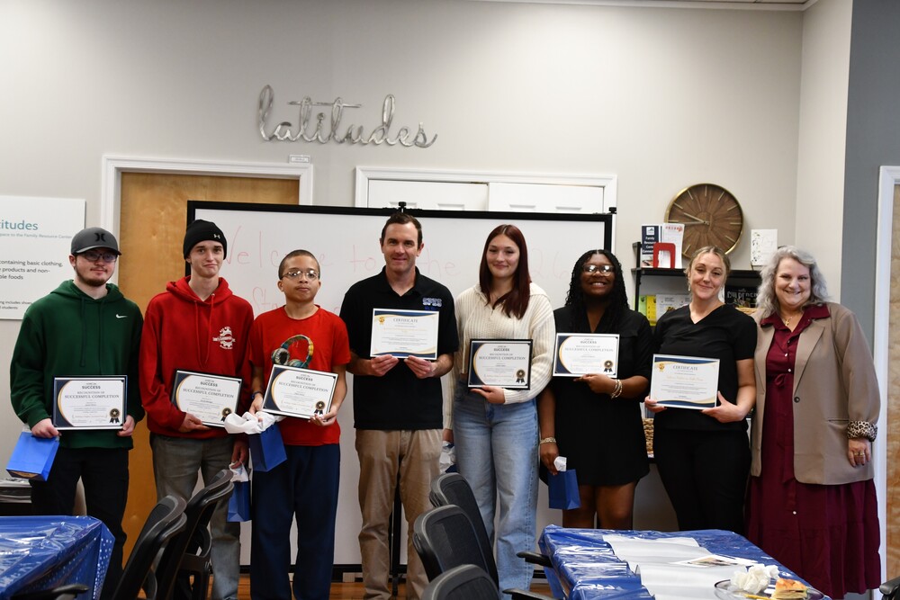 group  of students and adults standing with certificates under a silver sign on the wall that says latitudes