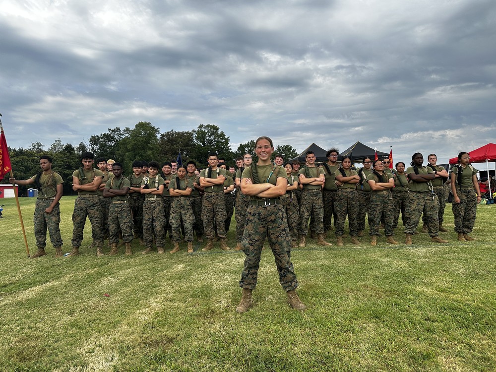 MCJROTC Raider Team poses in formation on a grassy field. One cadet stands confidently in front with arms crossed, while the rest of the team stands behind in rows, also with arms crossed. All cadets are in camouflage pants and olive green shirts, with tents and trees in the background under a cloudy sky.