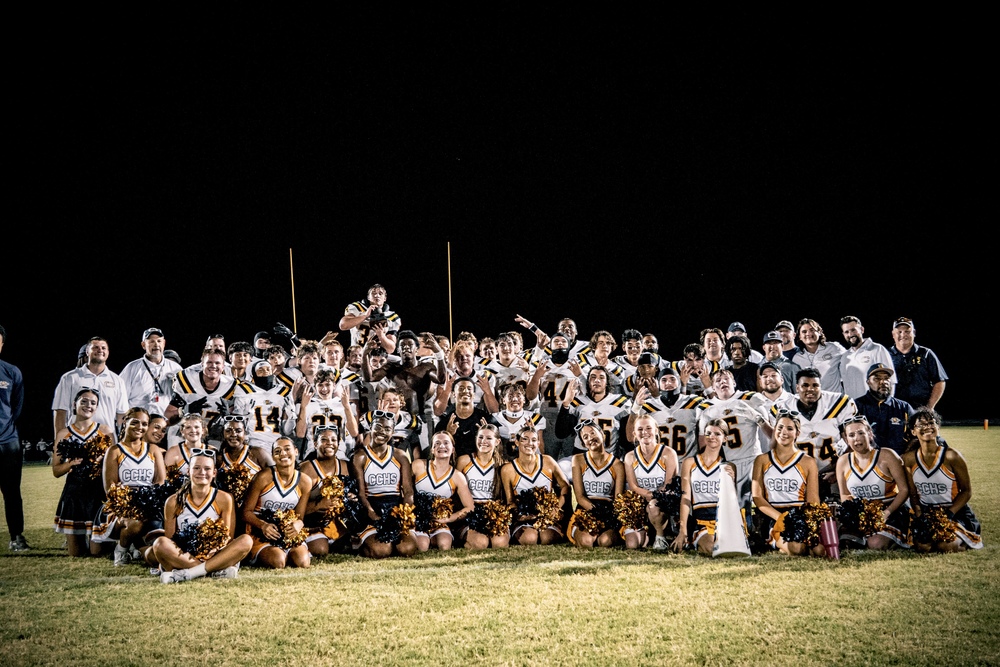The Culpeper County High School football team and cheerleaders celebrate together on the field at night after their historic Cannonball Classic victory. Players in white uniforms with black and yellow accents pose behind cheerleaders in navy and gold uniforms, all smiling and cheering with coaches and staff gathered around them.