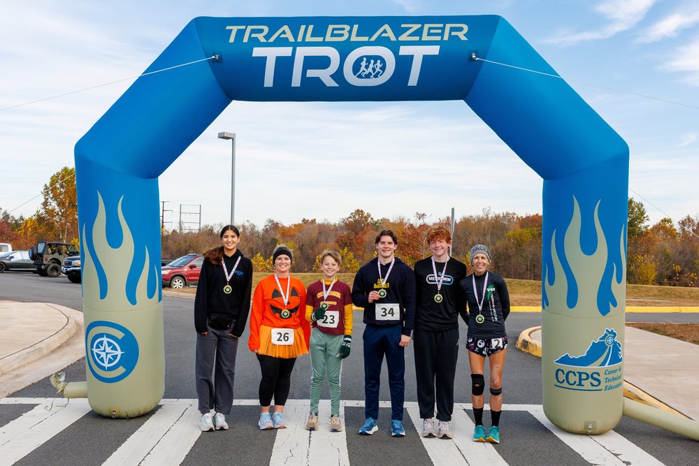 6 people wearing medals standing under a gold and teal blow up finish line arch that says trailblazer trot across the top