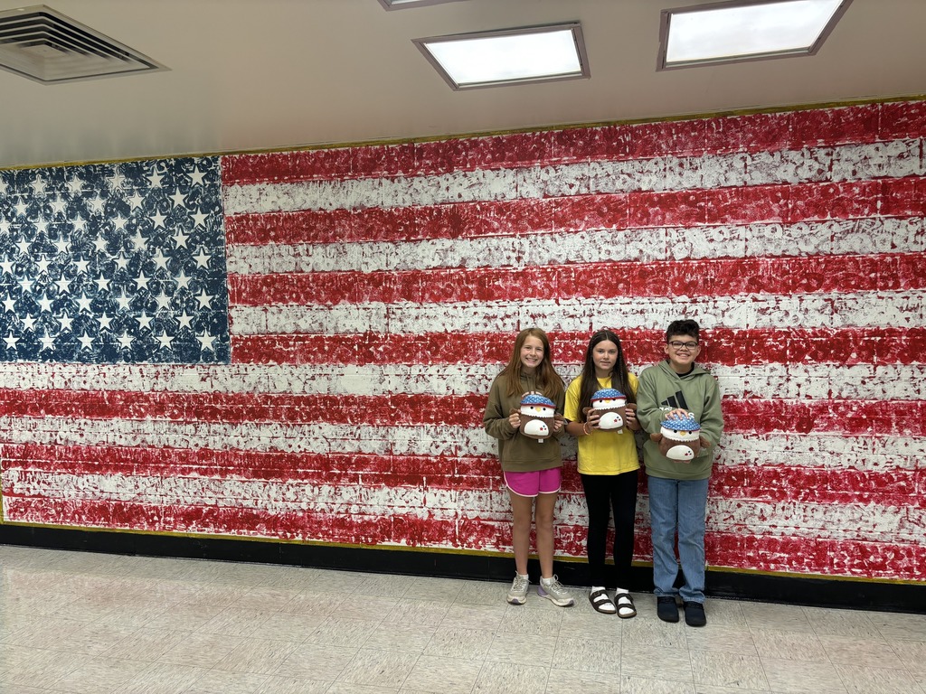 Three students stand in front of a large painted American flag mural, each holding a patriotic plushie. The students are smiling and dressed casually, displaying their prizes for a contest.