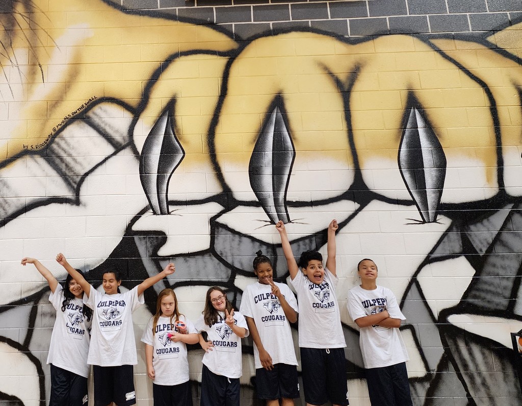 It’s a photo of the Cougar Champions team posing in front of a mural that shows a large, stylized cougar claw. The students are all wearing matching "Culpeper Cougars" shirts, and they’re joyfully raising their arms and smiling. The mural adds a fierce, energetic vibe to the scene.