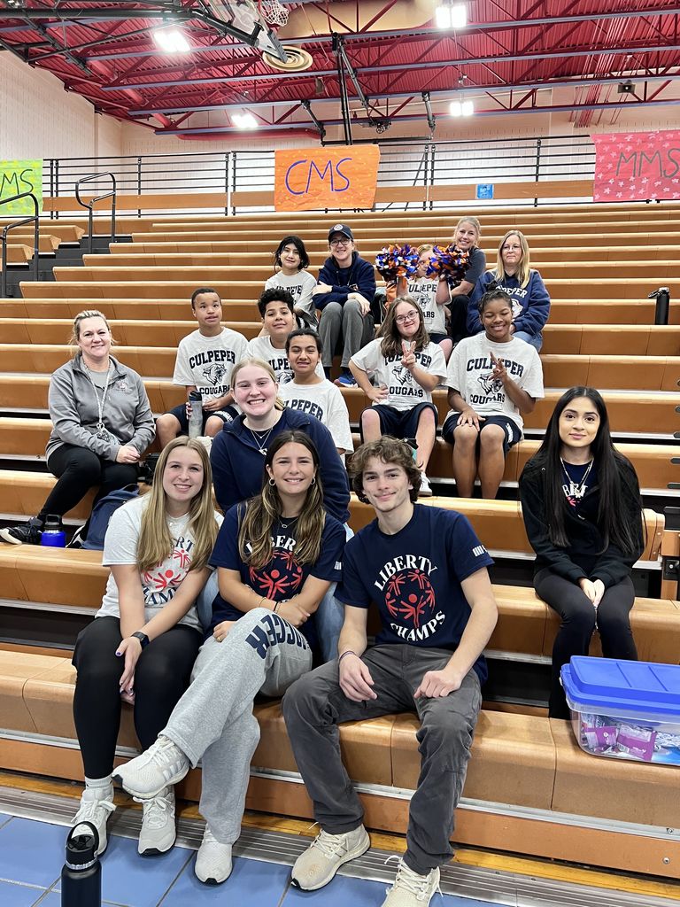 This image shows the Cougar Champions team sitting together on bleachers, joined by coaches and buddies from Liberty High School. The students are wearing their "Culpeper Cougars" shirts, while some Liberty students wear "Liberty Champs" shirts. The group looks relaxed and happy, with some holding pom-poms, showing team spirit and camaraderie. The gym setting includes banners with school names, adding to the event atmosphere.