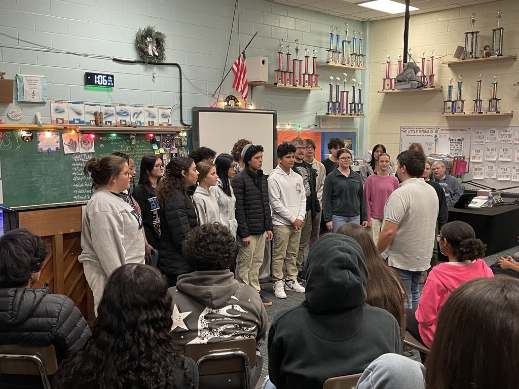 A high school choir is performing in a classroom, facing a group of middle school students seated in chairs. The choir consists of about a dozen students, with their teacher standing in front of them, addressing the group. The room is decorated with trophies on shelves, a chalkboard with musical notes and classroom rules, and festive string lights. The audience is engaged, watching the performance attentively.