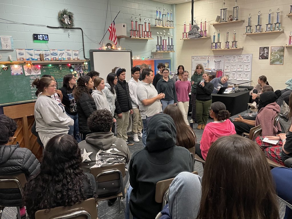 A high school choir stands in a classroom, engaging with a seated group of middle school students. The choir is composed of students in casual attire, with their teacher speaking to the audience. The classroom is decorated with trophies on shelves, a chalkboard with music-related content, and string lights. Students in the audience are seated in rows, attentively watching and listening to the interaction. The setting conveys an atmosphere of learning and musical appreciation.
