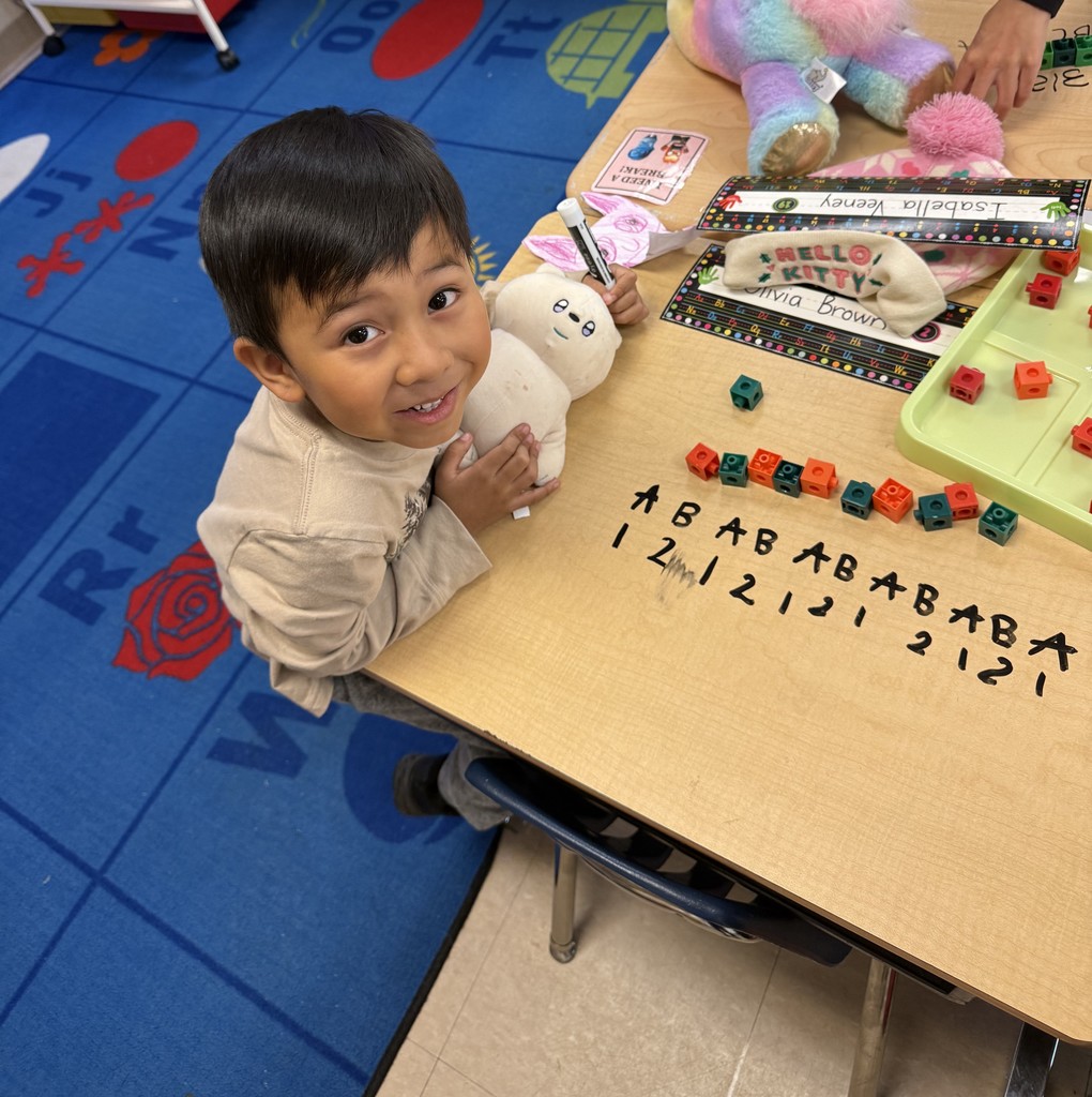 A smiling young boy sitting at a desk, holding a plush toy. On the desk are colorful blocks and written patterns, including "ABABAB" and numbers like "2121."