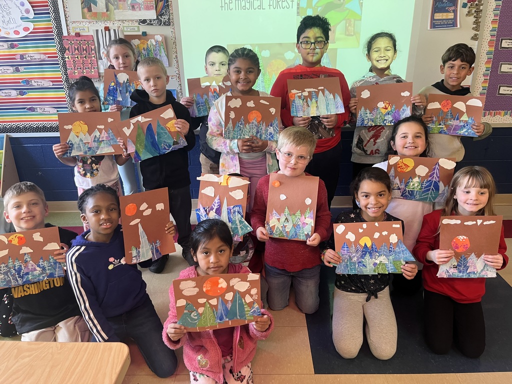 "A group of 15 children standing and kneeling in a classroom, holding up colorful art projects depicting snowy forests with mountains, trees, and suns or moons. The children are smiling and standing in front of a decorated classroom wall with posters and bulletin boards. The art projects feature a mix of brown, blue, green, and orange tones, with cut-out shapes and white clouds."