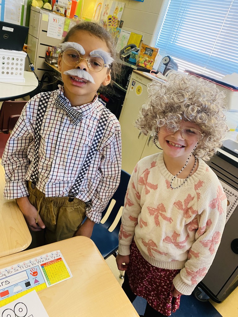wo children dressed as elderly individuals for a "100 Days of School" celebration. The child on the left wears a plaid shirt with suspenders, a bowtie, glasses, and a fake mustache and eyebrows. The child on the right has curly gray hair, glasses, and a sweater with pink bows, paired with a patterned skirt. They are standing in a brightly lit classroom near desks and colorful decorations.