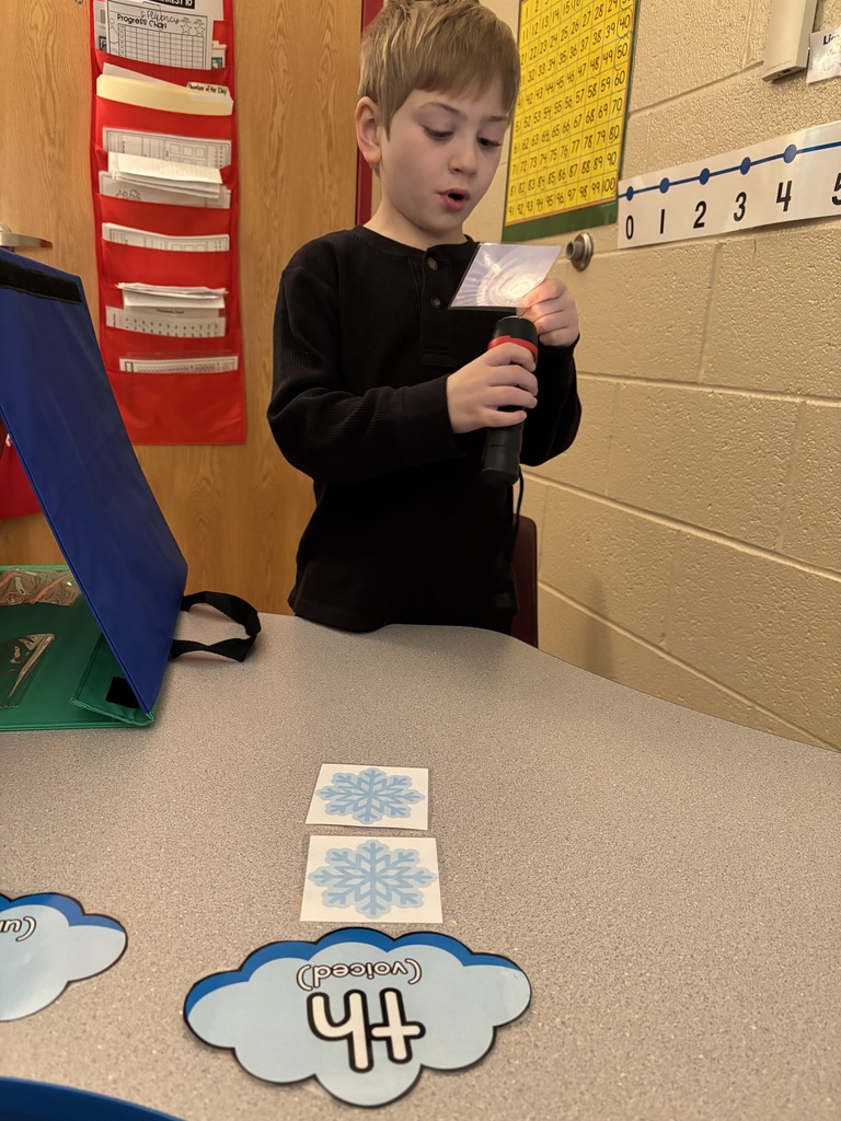 A young boy with short light brown hair, wearing a black long-sleeved shirt, is holding a flashlight and looking at a transparent card. The table has laminated snowflake cards and a word cloud card with "th" written on it. The background includes a red hanging organizer on a door and a number chart on the wall.