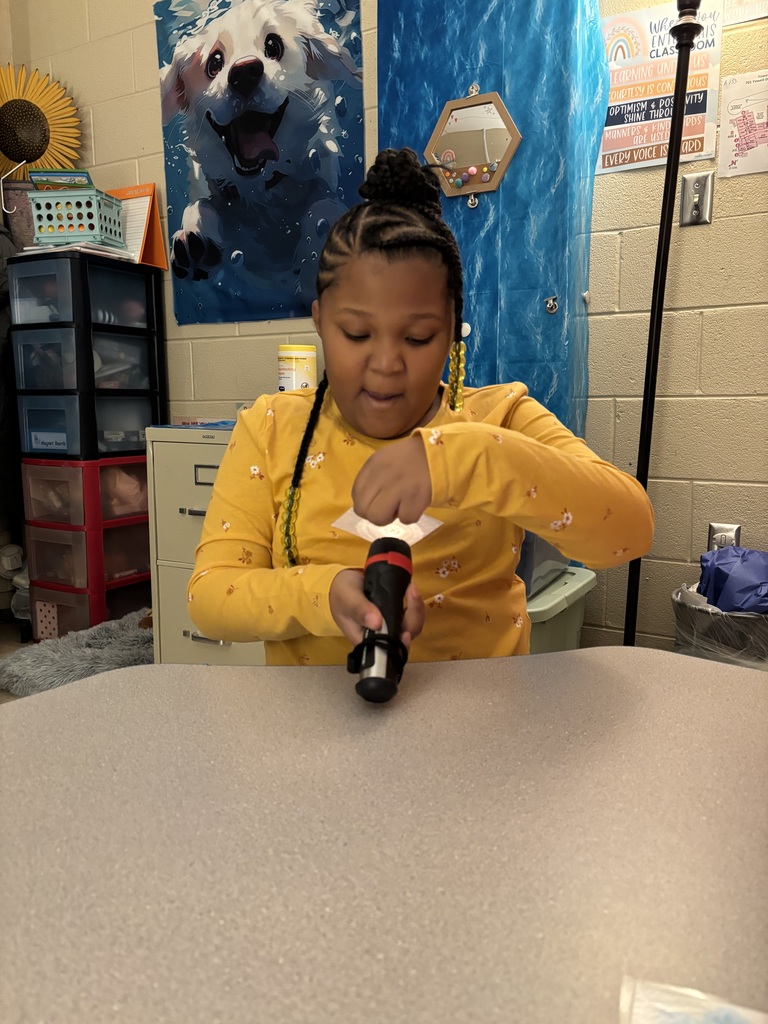 A young girl with braided hair, wearing a yellow long-sleeved shirt, is holding a flashlight and shining it on a piece of paper. She is seated at a classroom table with a blue background decoration behind her, along with a poster that has motivational words.
