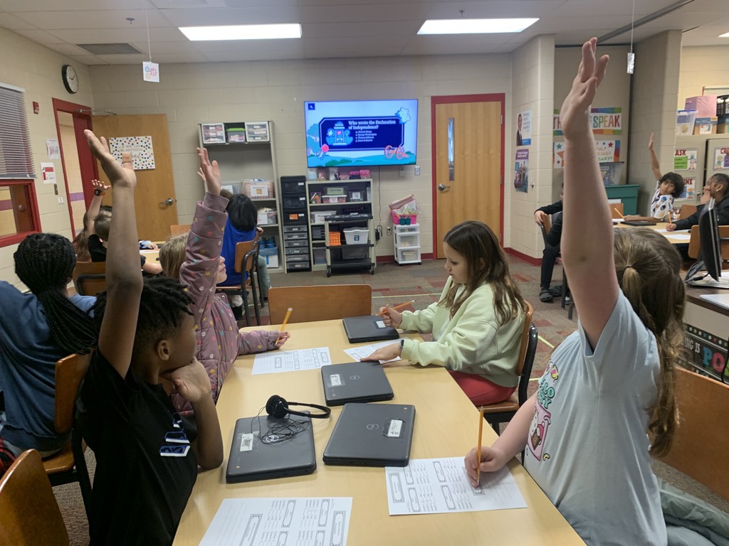 A classroom of elementary students with their hands raised, participating in a lesson. Laptops and worksheets are visible on the desks. The large screen at the front of the room displays an educational question related to history.