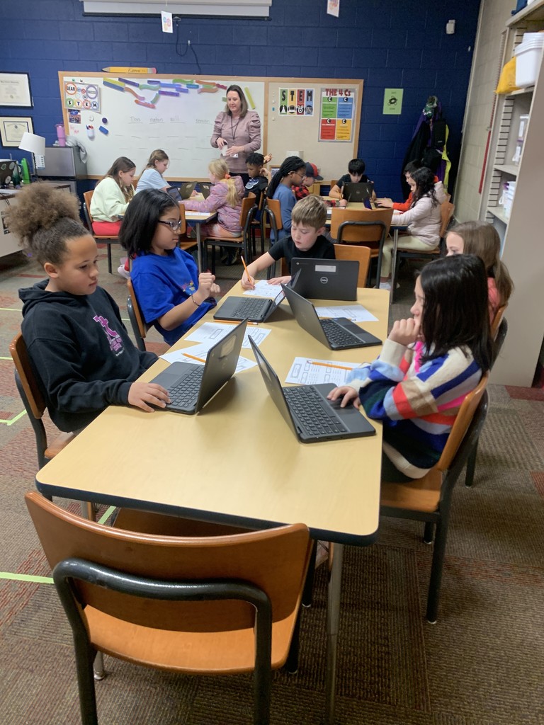 A classroom of elementary students engaged in a lesson while using laptops and worksheets. The teacher stands at the front near a whiteboard with STEM-related posters. Students appear focused, collaborating in small groups.