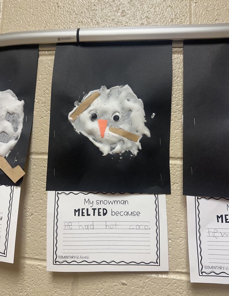 A classroom display of student artwork features a craft of a melted snowman made from shaving cream, googly eyes, a small orange paper carrot nose, and brown paper arms. Below it, a student has written, "My snowman melted because he had hot cocoa."