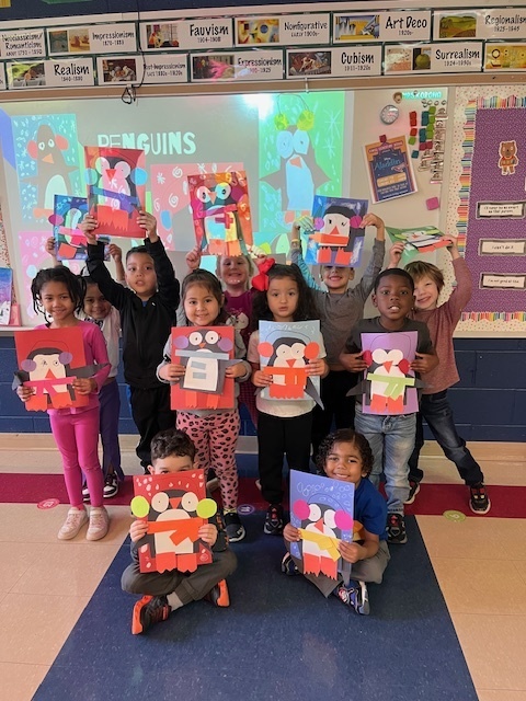 A group of young students in a colorful classroom proudly holding up their handmade penguin crafts. The crafts are made from cut-out paper in various colors, with some featuring heart shapes. The classroom walls are decorated with educational posters and artwork, and a projector screen in the background displays an image of penguins. The children are smiling and excited, showcasing their creativity.