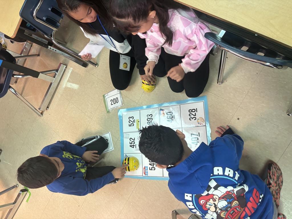Four children are collaborating around a floor grid with numbers and educational icons. They are programming Bee-Bots to navigate the grid, using number cards as prompts. The group is deeply engaged in computational thinking and strategy.
