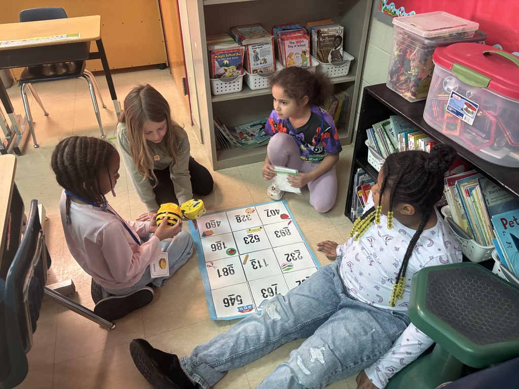 Four young students are positioned near a bookshelf, gathered around a grid mat with Bee-Bots. They are discussing strategies and taking turns programming the bots, practicing problem-solving and collaborative skills.