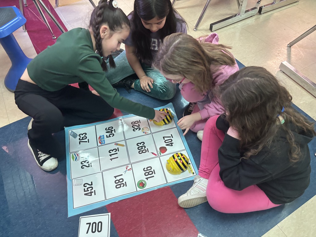 Four elementary school students are gathered around a colorful grid mat on the floor, programming yellow and black Bee-Bots to navigate between numbered squares with educational icons. One student points to a square, actively engaging in problem-solving with her peers.