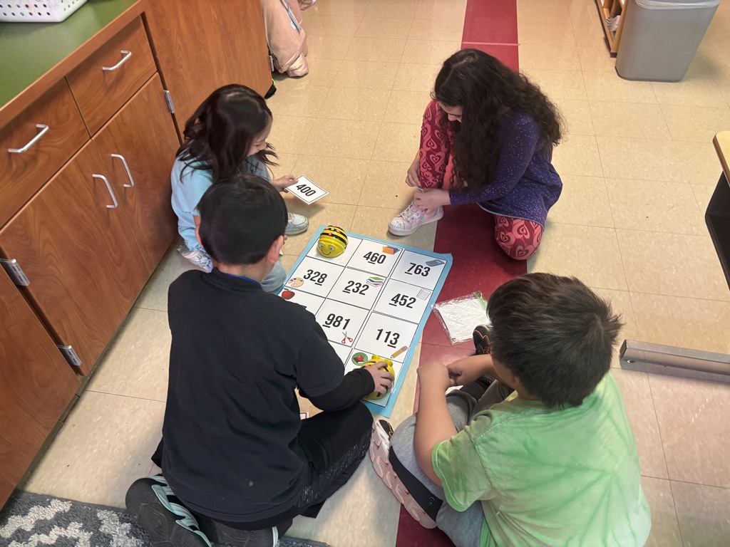 Four students are sitting on the floor next to a cabinet, working together on a numbered grid mat with Bee-Bots. One child is holding a number card while another guides a Bee-Bot, emphasizing cooperative learning and coding skills.