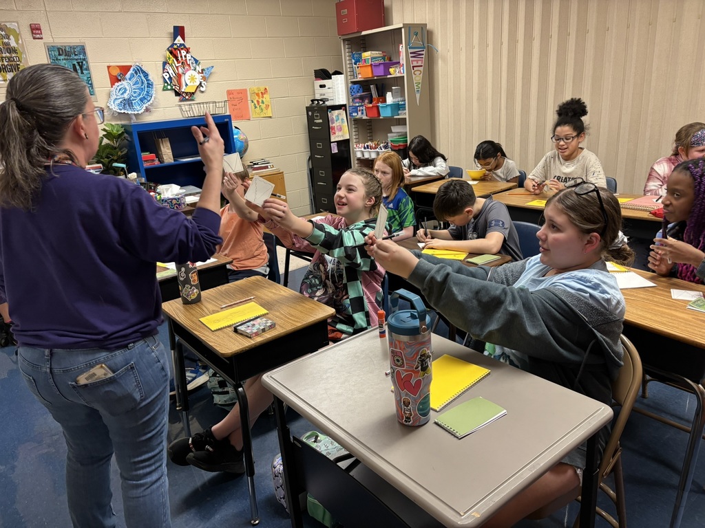 a group of excited students hold their sketched up for their teacher to see. The teacher has her arms raised in enjoyment of their creations. the students are seated at desks and smiling.