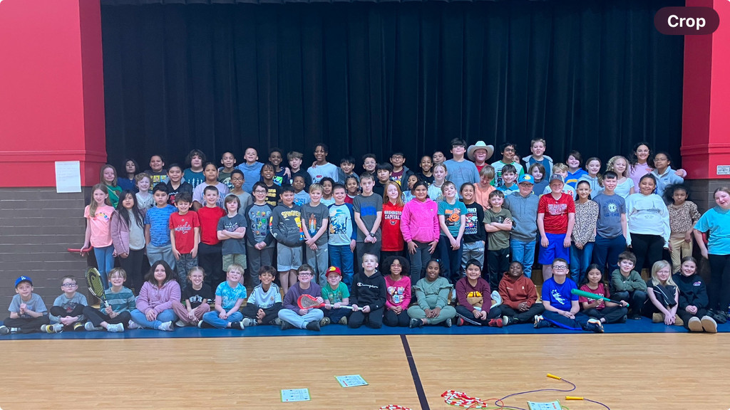 A larger group of elementary school students gathers for a group photo in the same gymnasium setting, in front of a black stage curtain. The students are dressed in a mix of casual clothing, including some wearing hats. They are arranged in several rows, with some sitting on the floor and others standing on the risers. A few students are holding sports items such as a tennis racket and jump ropes, which are also scattered on the floor in the foreground. The gymnasium has red and brick walls.