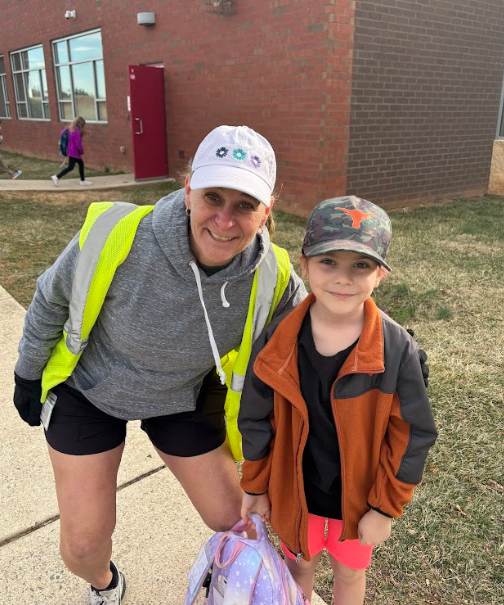 A woman wearing a white baseball cap with floral designs, a gray hoodie, black shorts, and a reflective safety vest is smiling next to a young boy in a camouflage baseball cap, an orange and black jacket, and pink shorts. They are standing outside a brick school building with a red door in the background. The boy is holding a pastel-colored backpack, and other children can be seen walking behind them.