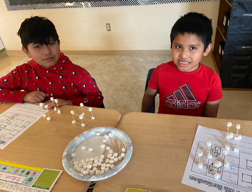 Two boys sitting at a desk in a classroom, working on a STEM activity. They are using marshmallows and toothpicks to construct geometric structures. A plate filled with marshmallows and toothpicks sits between them. Both boys are smiling, and a worksheet is visible on the desk.