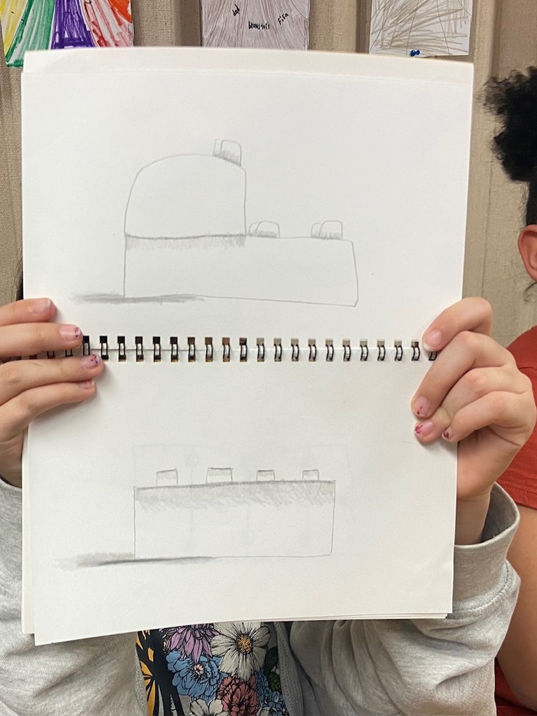 A student holds up a sketchbook displaying two pencil drawings of simple block-like structures with rounded elements on top. The drawings incorporate shading to add depth. The student’s hands, with slightly painted fingernails, are visible, along with a portion of their floral-patterned clothing.