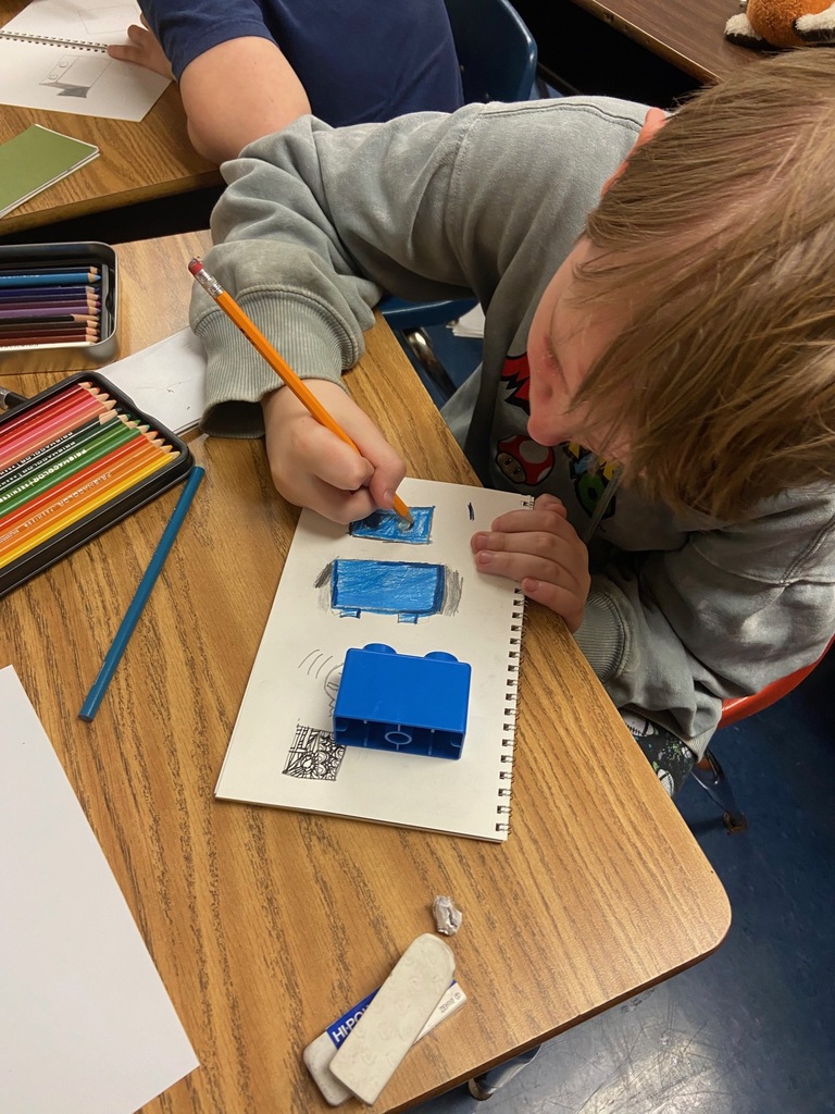 A student in a gray hoodie colors a blue building block drawing in their sketchbook with a colored pencil. The actual blue block is placed on the paper for reference. The desk is scattered with art supplies, including colored pencils, an eraser, and a pencil sharpener.