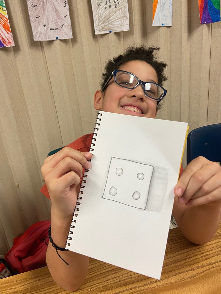 A smiling student with curly hair and glasses holds up a spiral sketchbook, displaying a pencil drawing of a die. The drawing includes shading to create a 3D effect. The student sits at a desk in a classroom, with colorful student artwork pinned on the wall behind them.