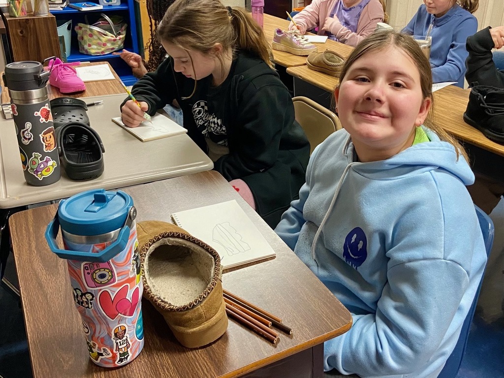 A smiling student in a light blue hoodie sits at a desk with a brown slipper in front of them. Their sketchbook contains a partially drawn shoe. Other students are seen in the background, focused on drawing different types of shoes.