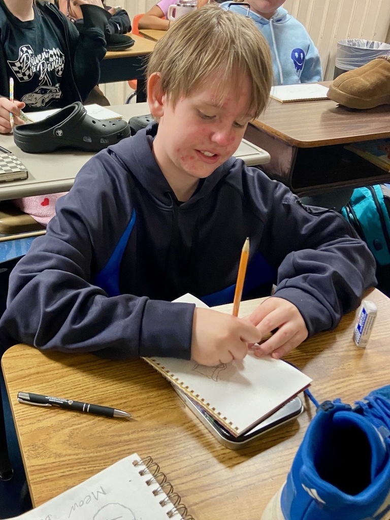 A student with short blond hair, wearing a navy blue hoodie, works on a shoe sketch in a spiral notebook. Other students are seen in the background, sketching their own shoes, with various sneakers and slippers placed on desks.