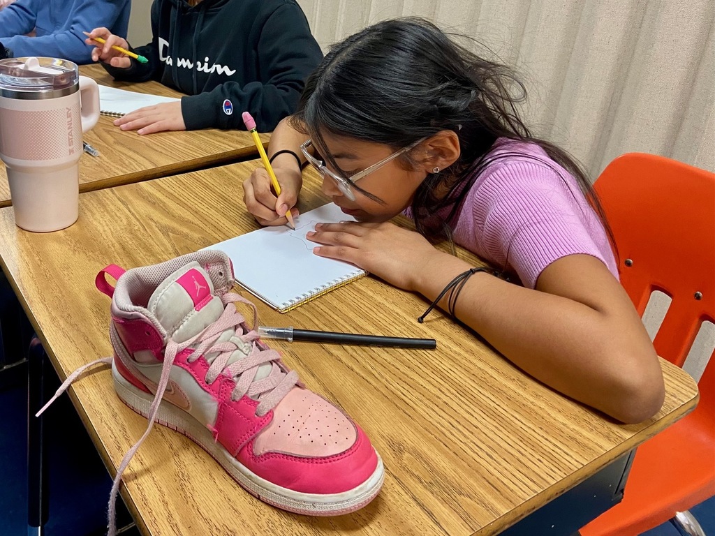 A student wearing glasses and a pink shirt leans over their sketchbook, concentrating on drawing a pink and white high-top sneaker that is placed on the desk. The student holds a yellow pencil, and a large pink tumbler sits nearby.