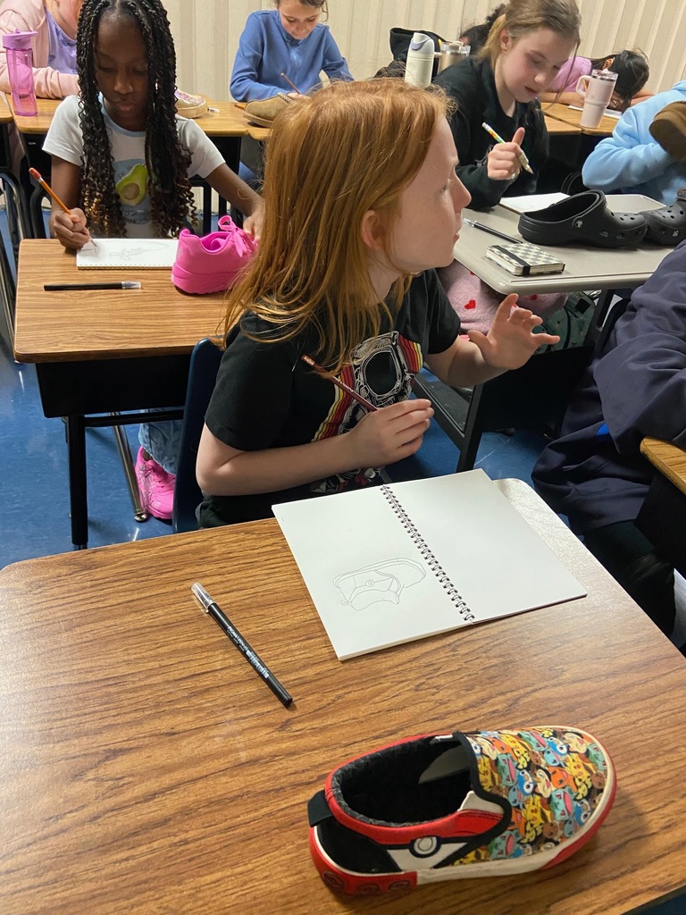 A red-haired student in a black t-shirt is mid-conversation while sitting at a desk with a spiral sketchbook open to a shoe drawing. A colorful Pokémon-themed slip-on shoe sits on the desk. Another student behind them is also sketching a bright pink sneaker.
