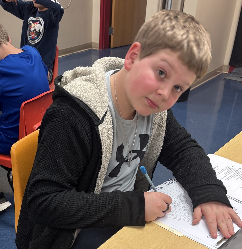 A student in a hoodie and Under Armour shirt is working at a desk, writing on a worksheet. He looks up from his work, glancing toward the camera.